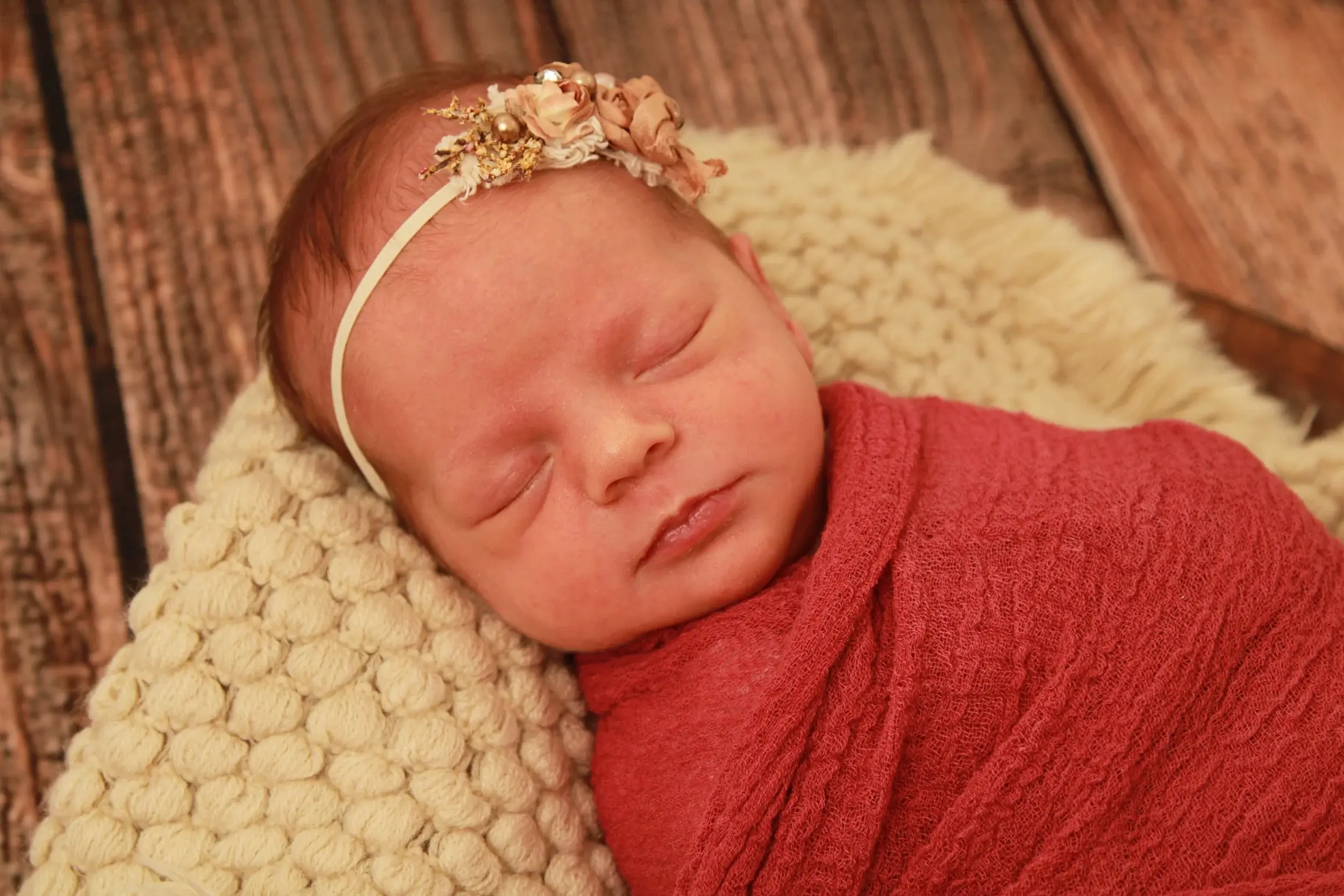 Newborn baby girl wrapped in a red knit wrap, photographed against a rustic wood backdrop — serving Atlantic Beach, NY families